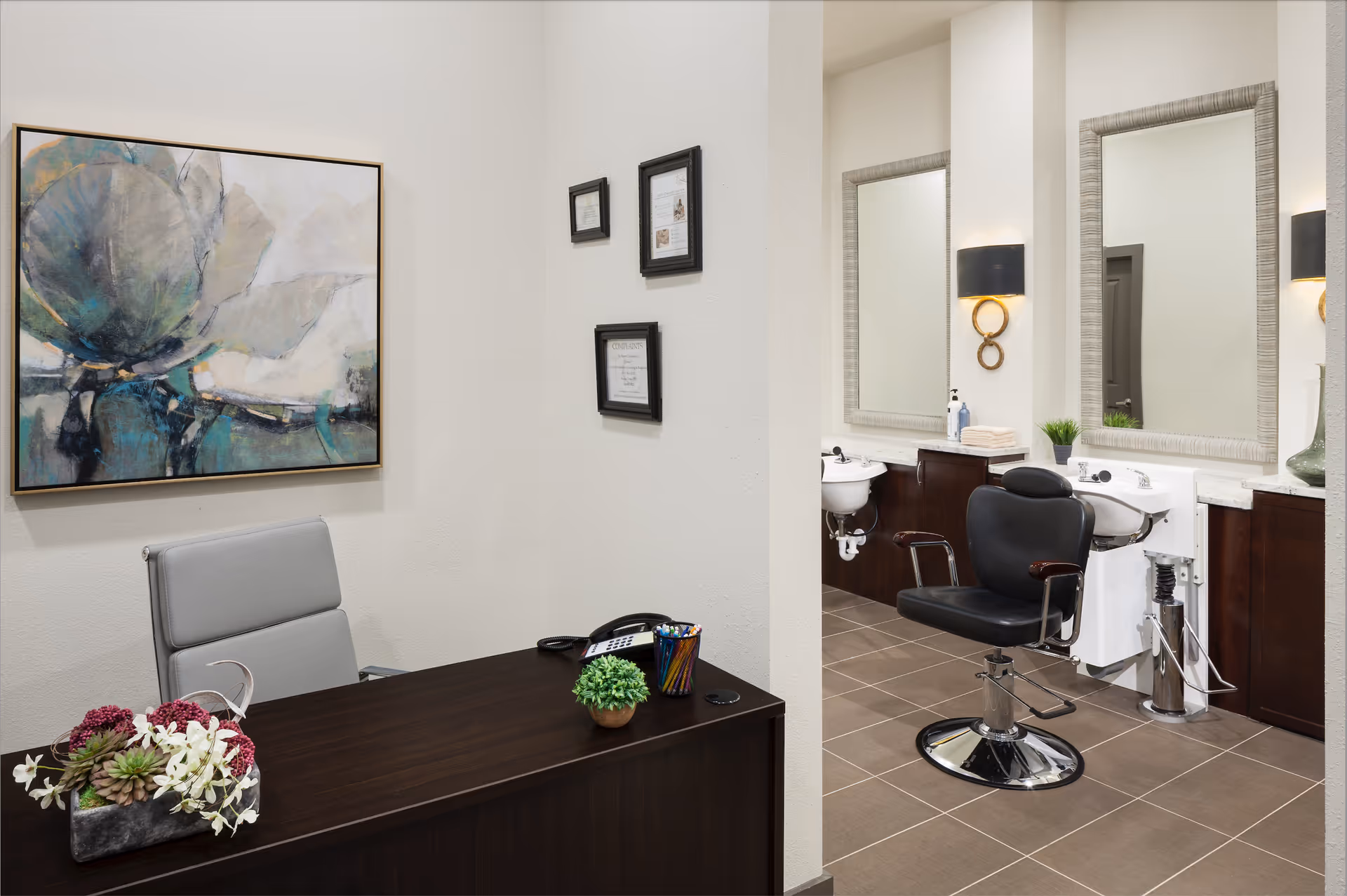 Interior view of a salon area within a senior living facility. The foreground shows a dark wooden desk with a gray office chair, a small plant, a phone, and a container with colored pencils. On the wall behind the desk is a large abstract painting. In the background, there is a salon chair in front of a sink and two large mirrors with wall sconces on either side. The floor is tiled in a neutral color.