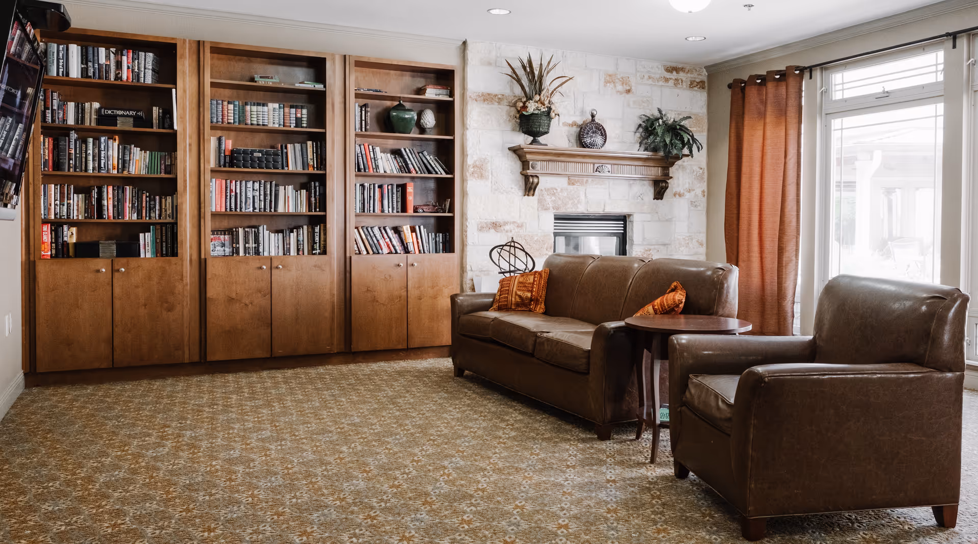 A cozy living room with a brown leather sofa and armchair, a wooden side table, a stone fireplace with decorative plants on the mantle, and built-in wooden bookshelves filled with books. Large windows with brown curtains allow natural light to enter the room.