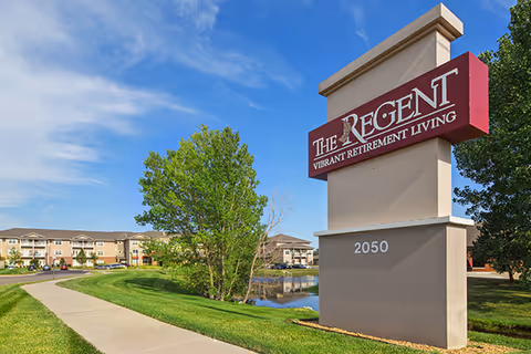 Outdoor view of The Regent retirement living facility sign with a clear blue sky, green grass, a sidewalk, trees, and the building in the background.