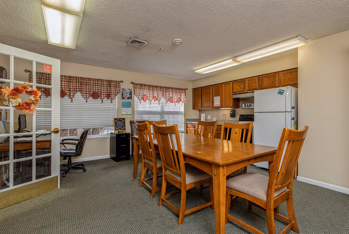 Communal dining area with a wooden table and chairs, a kitchenette with refrigerator and cabinets, and windows with patterned valances.