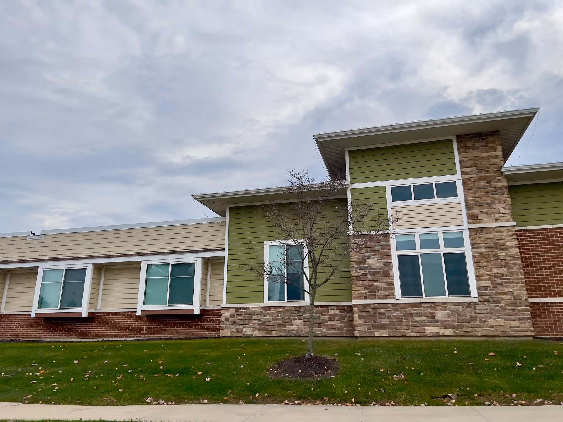 Exterior front view of a low-rise building with green and beige siding, stone accents, multiple windows, and a small tree on a grassy lawn.