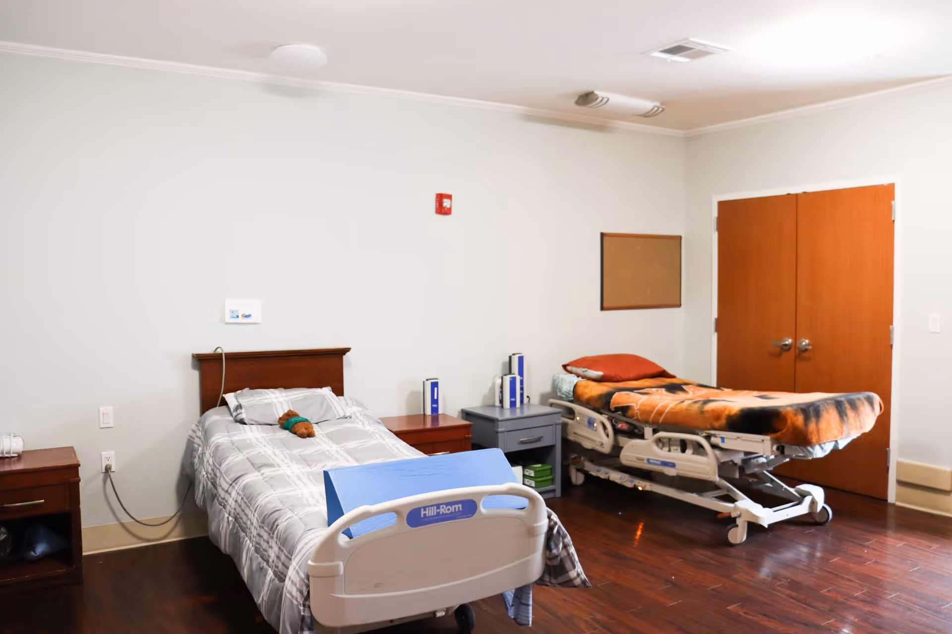 A room with two hospital-style beds, one with a gray and white checkered blanket and a small stuffed toy on it, the other with an animal print blanket and a red pillow. There are two wooden nightstands between and beside the beds, a corkboard on the wall, and double wooden doors in the background. The room has light-colored walls and dark wooden flooring.