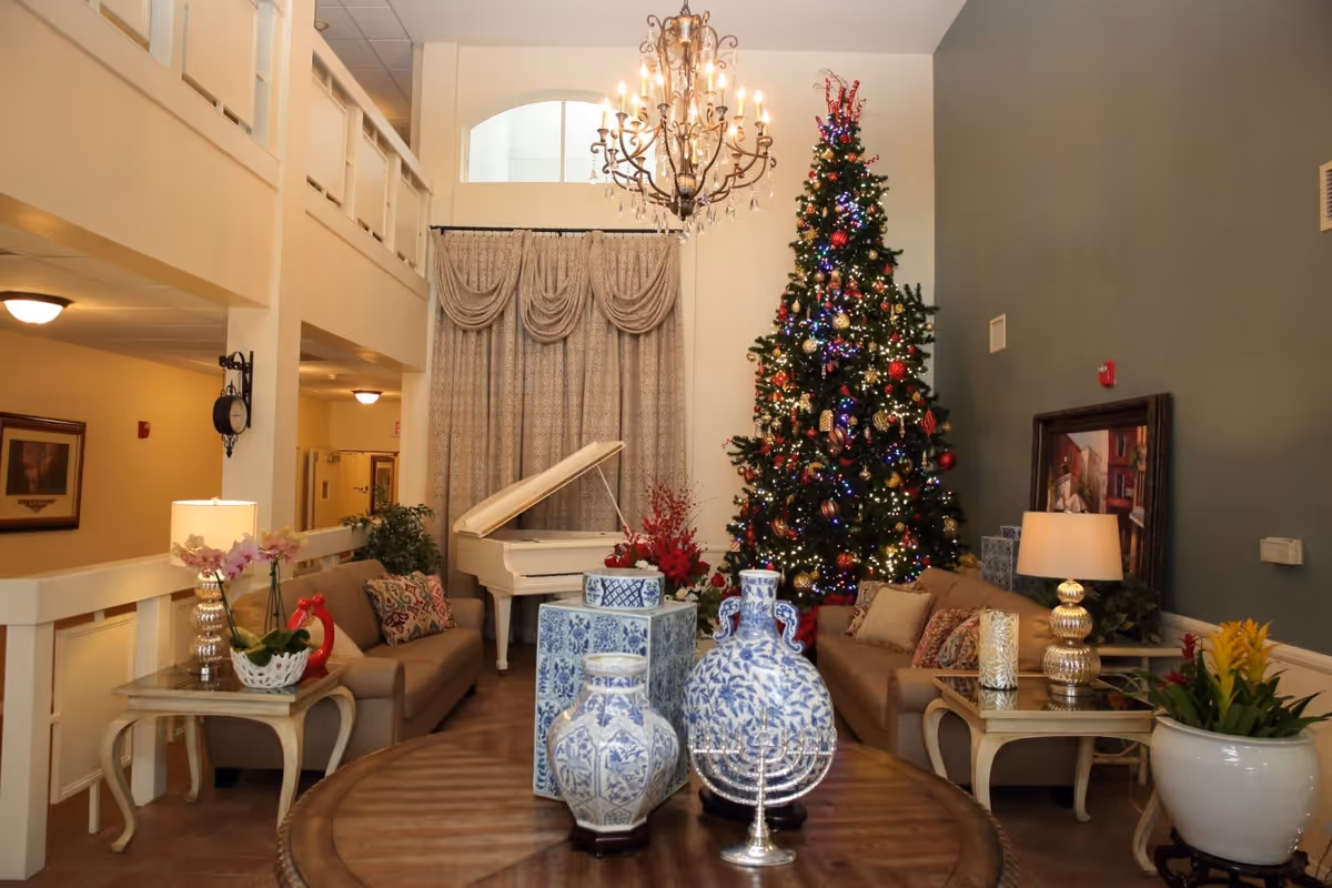 A cozy senior living facility common area decorated for the holidays with a tall Christmas tree adorned with lights and ornaments. The room features two beige sofas with patterned pillows, a white grand piano, a chandelier, and a round wooden table with blue and white decorative vases. There are also side tables with lamps and plants, and a framed painting on the wall.