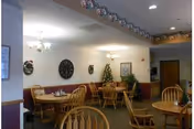 Dining area with several wooden tables and chairs arranged neatly. The room is decorated with a Christmas tree, wall clock, framed pictures, and holiday garlands hanging from the ceiling. The walls are light-colored with a wooden trim along the bottom.