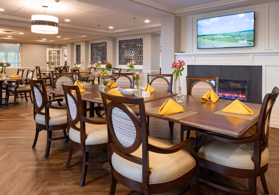 A dining room in a senior living facility with wooden tables and chairs. Each table is set with yellow folded napkins and small vases containing pink flowers. There is a modern electric fireplace beneath a wall-mounted TV displaying a nature scene. The room has warm lighting and decorative wall panels.