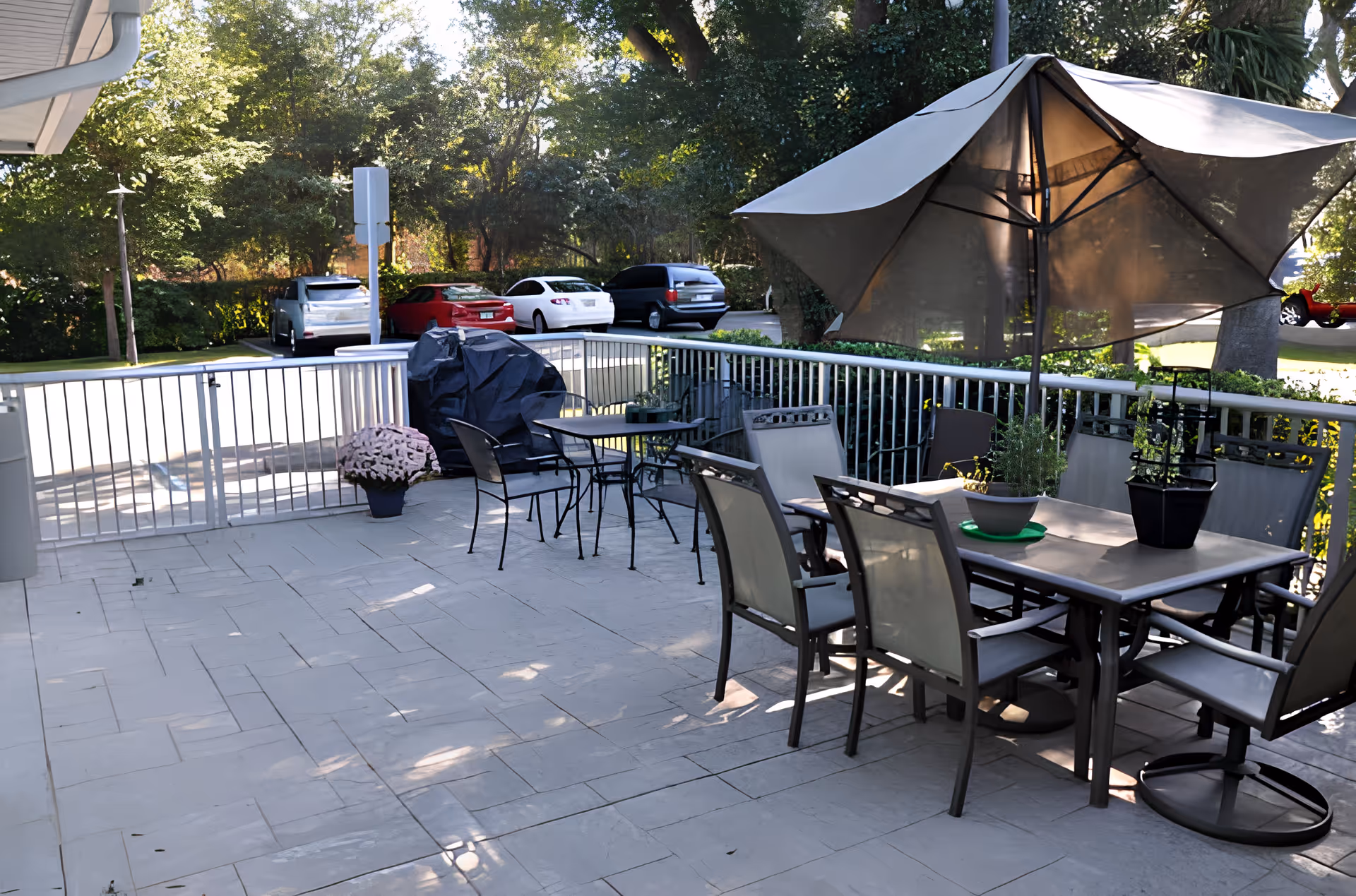 Outdoor patio area with a table and six chairs under a large umbrella, potted plants on the table, a smaller table with two chairs, a covered grill, and a parking lot with several cars and trees in the background.