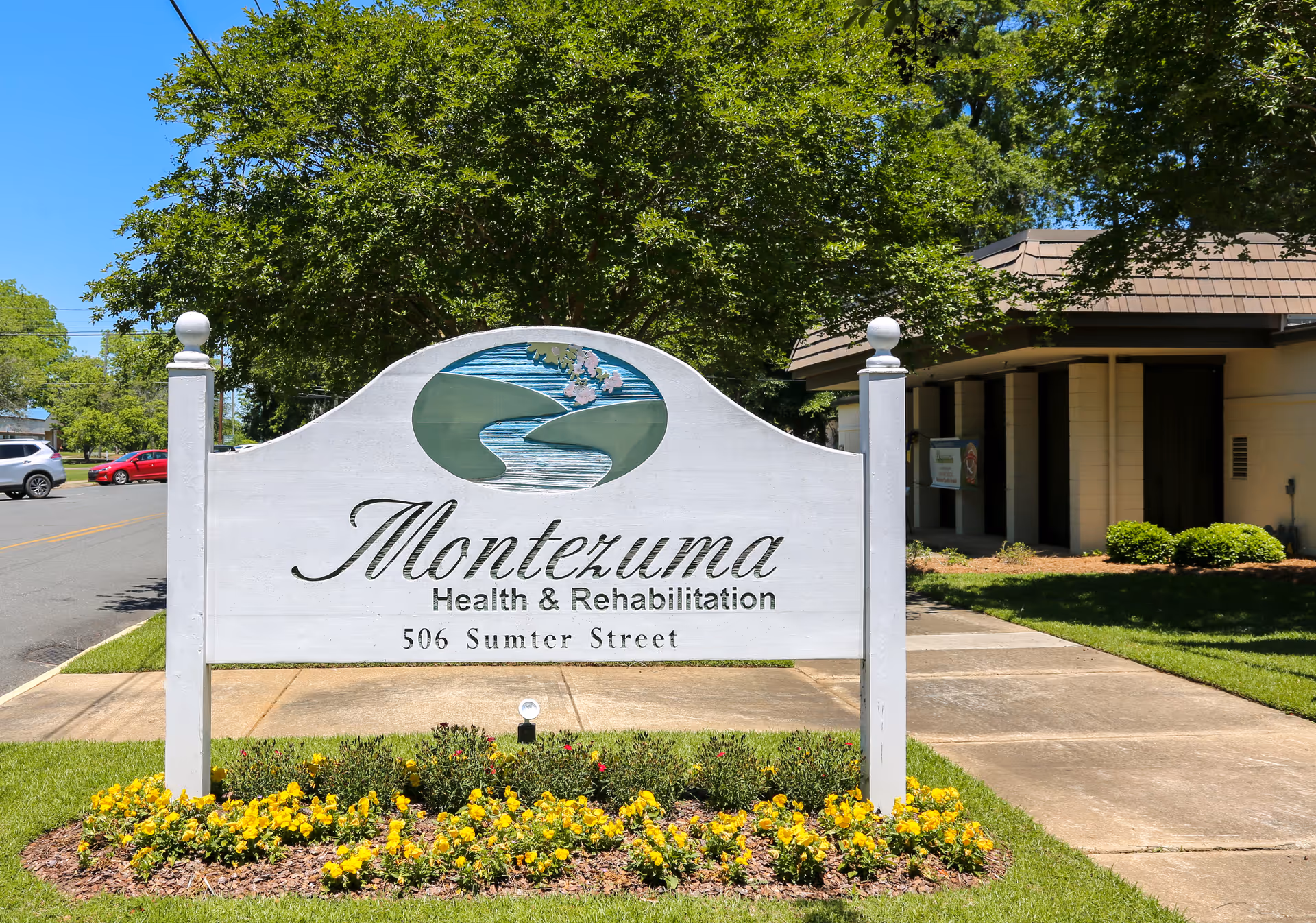 A white wooden sign reading 'Montezuma Health & Rehabilitation, 506 Sumter Street' stands in front of the facility with flowers, lawn, and the building entrance visible behind it.