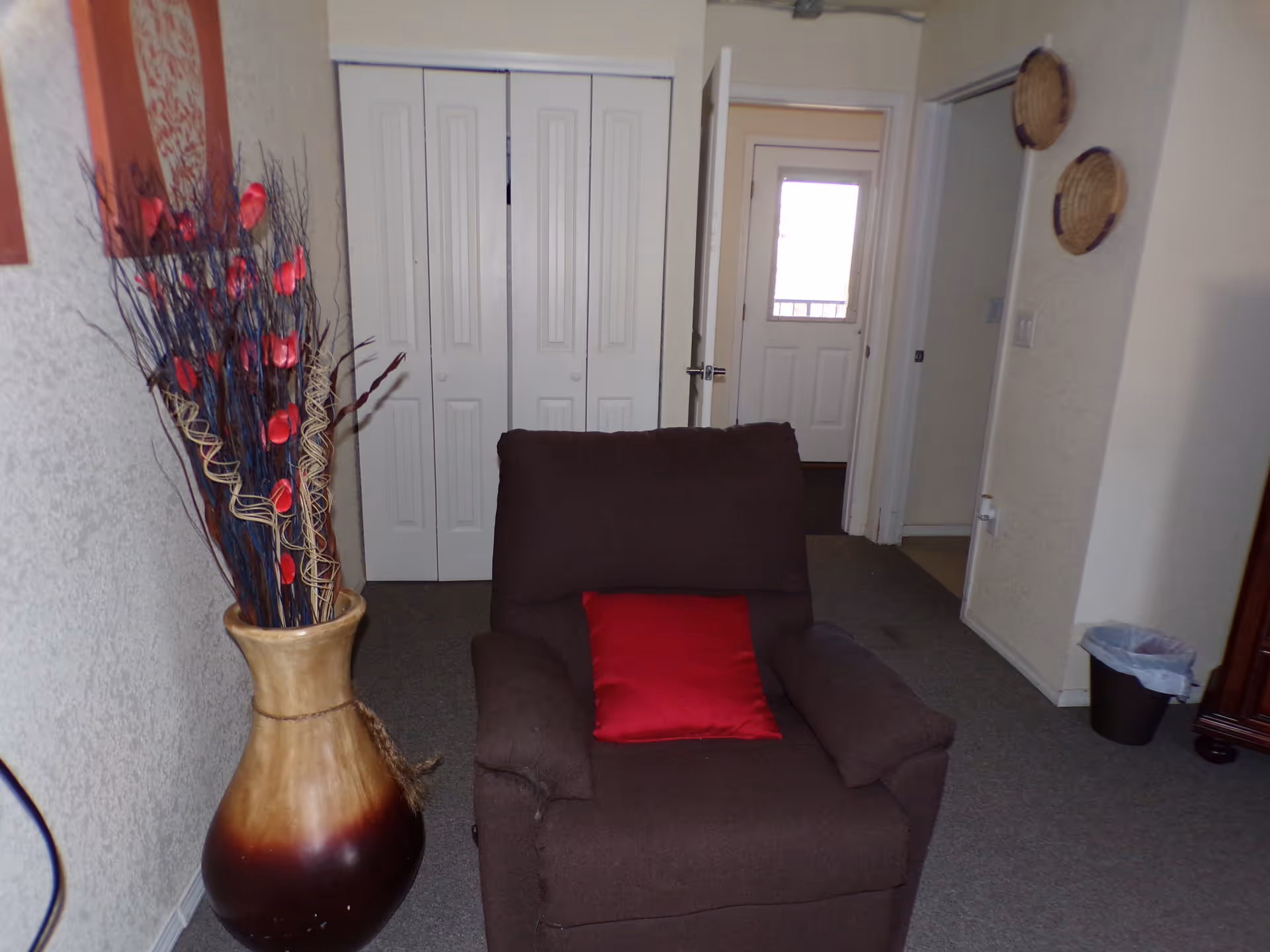A brown armchair with a red cushion placed in a carpeted room. To the left of the chair is a large decorative vase with tall dried branches and red flowers. Behind the chair, there is a white closet with double doors and an open door leading to another room with a white door that has a window. On the right wall, two woven baskets are hung, and a small trash bin is on the floor.