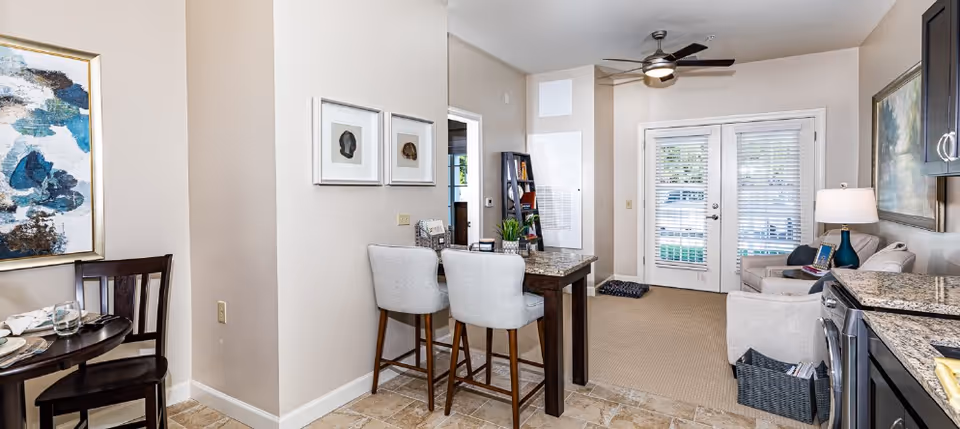 Interior view of a senior living facility showing a small dining area with a round table and two chairs on the left, a kitchen counter with granite countertops and appliances on the right, and a living room area with a sofa, armchair, lamp, and wall art in the background. There are double glass doors with blinds leading outside, and a ceiling fan above.