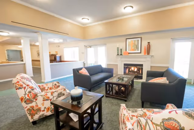Bright communal living room with sofas and floral armchairs arranged around a coffee table and a fireplace.