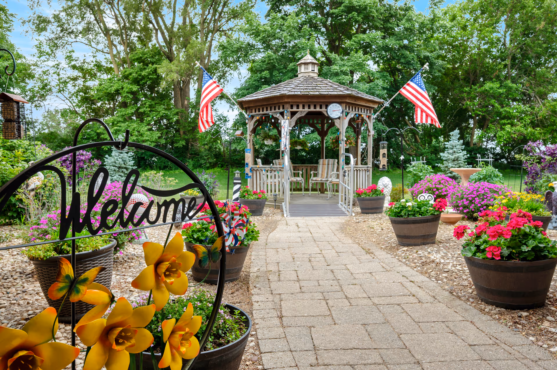 A garden walkway leading to a wooden gazebo surrounded by potted flowers, American flags, and a 'Welcome' sign.