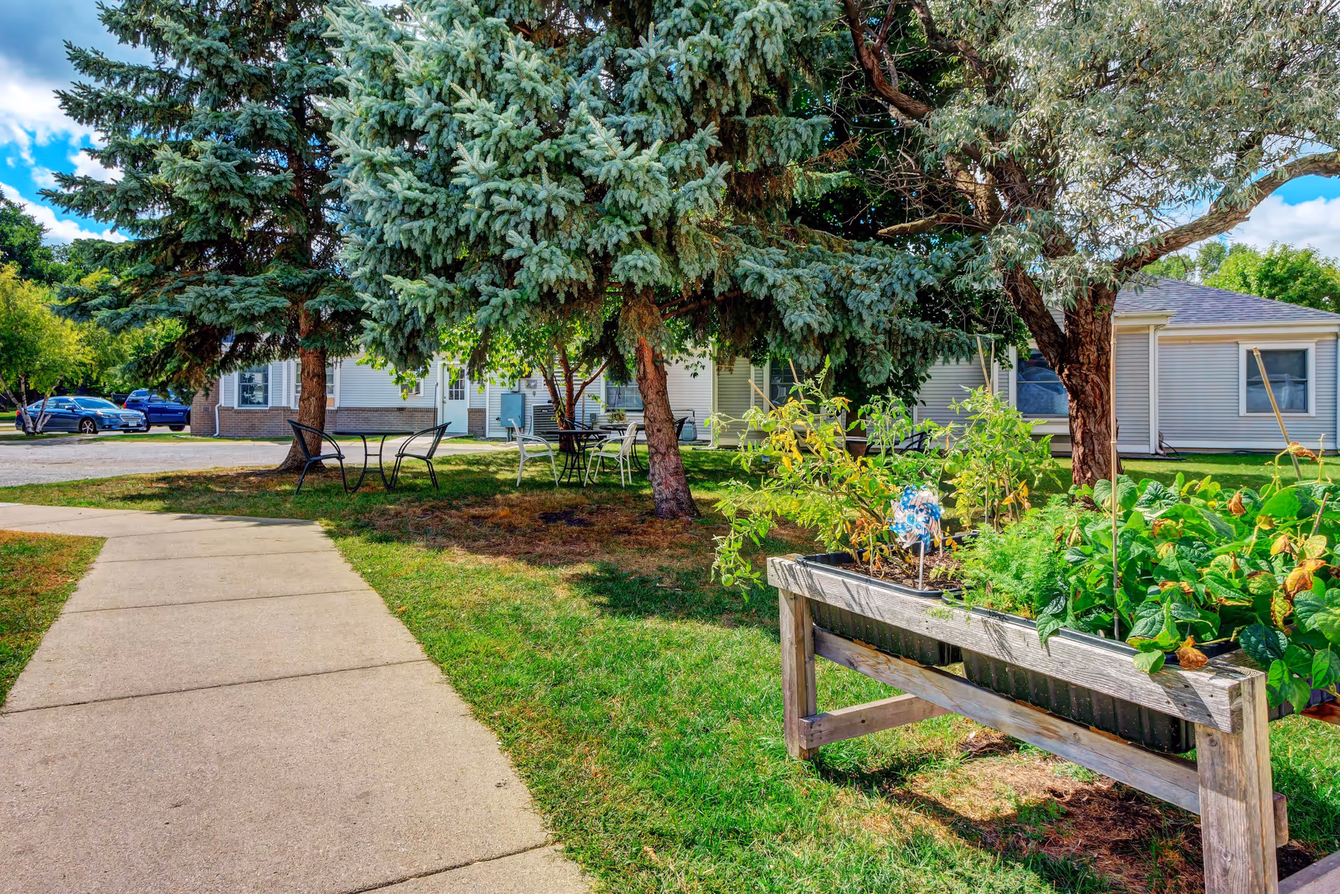 Outdoor garden area at Ecumen Evergreens Of Moorhead with a raised wooden planter box containing various green plants. There are large trees providing shade over metal tables and chairs on a grassy lawn. A paved walkway leads through the garden area towards a light-colored building in the background under a partly cloudy blue sky.