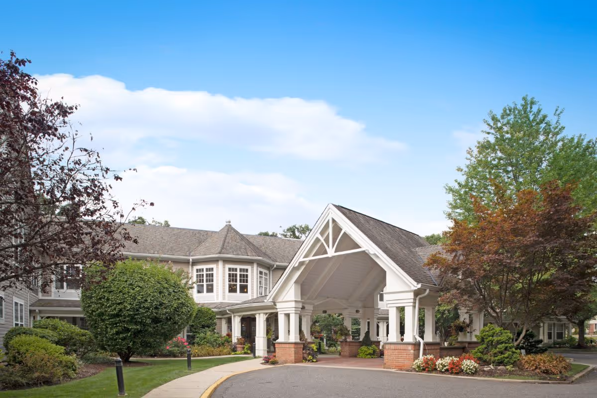 Entrance of a senior living building with a white porte-cochere, landscaped shrubs and a driveway under a blue sky.