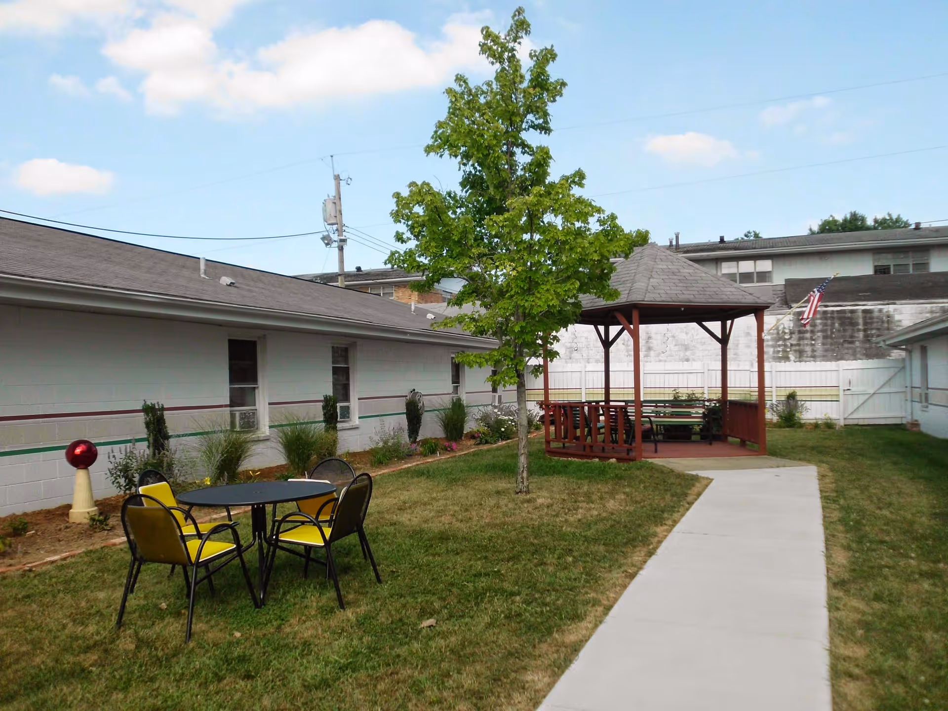 Outdoor area of Decatur Rehabilitation Health Care Center featuring a grassy lawn with a small tree, a round table with four chairs with yellow cushions, a wooden gazebo with benches, a concrete walkway, and buildings with white walls and windows surrounding the space under a partly cloudy sky.