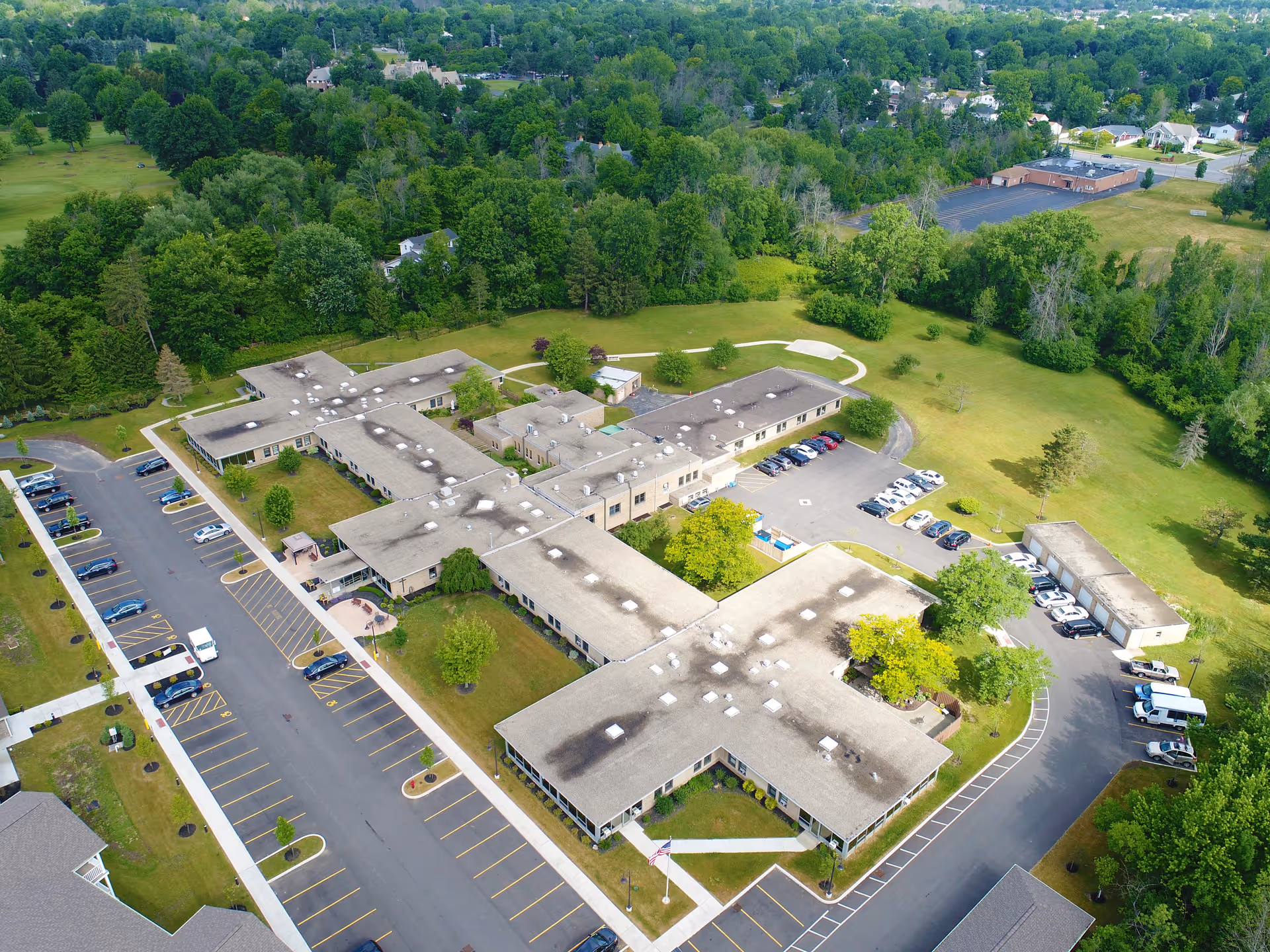 Aerial view of Park Creek Senior Living Community showing a large building complex with multiple wings surrounded by parking lots, green lawns, and trees. The facility is situated near a wooded area and residential neighborhood.