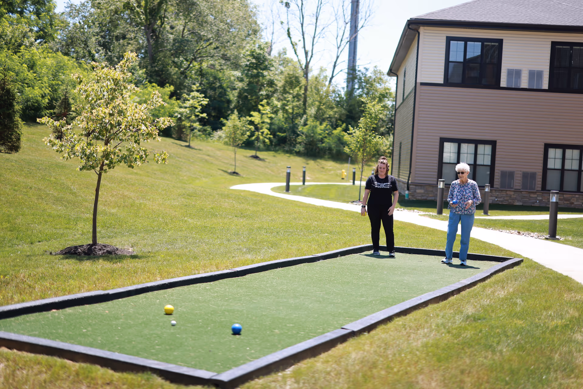Two women standing on a small outdoor bocce court on grassy grounds with a building in the background.