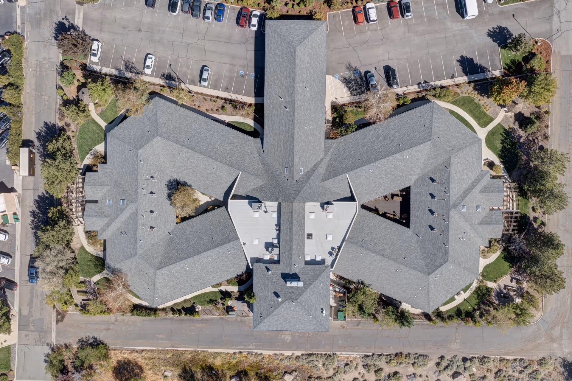 Aerial view of the Arbors Memory Care facility showing the building's roof layout, surrounding parking lots with cars, landscaped greenery, and pathways around the building.