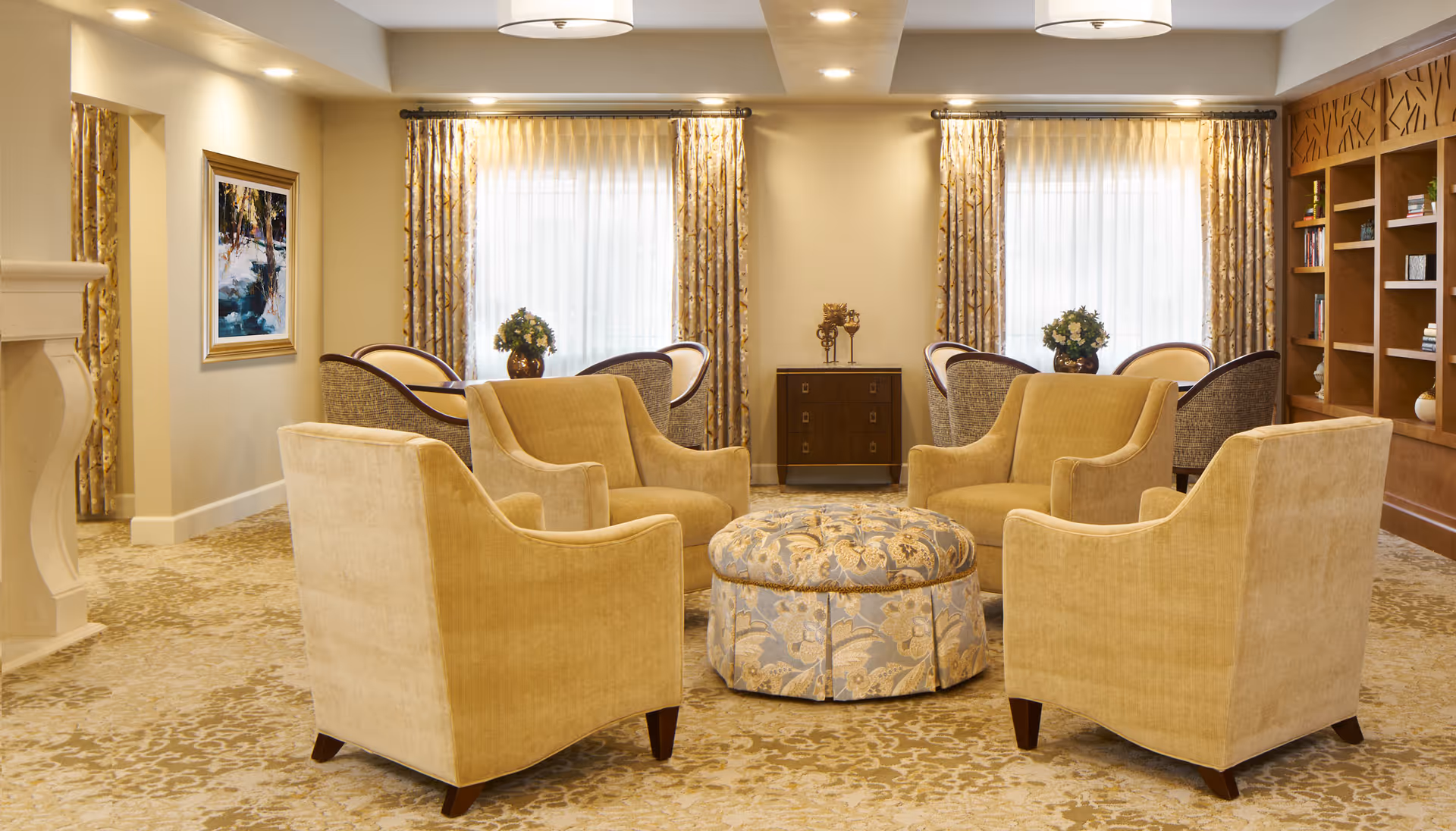 A cozy and elegant living room area with four beige armchairs arranged around a round patterned ottoman. Behind the chairs are two windows with sheer curtains and gold-patterned drapes. There are two small tables with flower arrangements and a wooden cabinet between the windows. On the right side, there is a built-in wooden bookshelf with books and decorative items. The room has warm lighting and a patterned carpet.