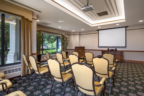 A small conference or presentation room with rows of beige cushioned chairs facing a wooden podium and a large white projection screen. The room has patterned carpet flooring, large windows with views of greenery outside, and recessed ceiling lights.