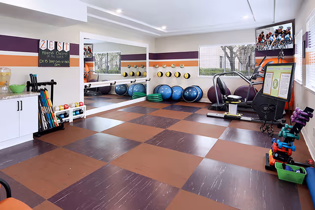 A fitness room with brown and maroon checkered flooring, exercise balls, dumbbells, resistance bands, and an exercise bike. The room has large windows with blinds, a wall mirror, and a TV mounted in the corner displaying a cycling race. There is a small counter with a water dispenser and a chalkboard sign that reads 'ZEST Fitness of the Week: Do 5 bicep curls or bicep arm'.