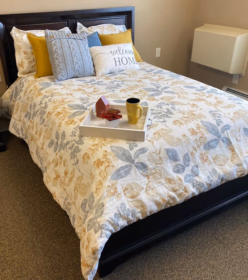 A neatly made bed with a floral patterned comforter in shades of beige, blue, and gray. The bed has multiple pillows including a gray knitted pillow, yellow pillows, and a white pillow with the words 'welcome HOME' printed on it. A white tray on the bed holds a small red potted plant, a small decorative house, and a yellow coffee mug. The room has beige walls and a carpeted floor.
