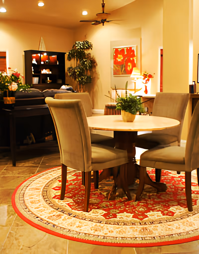 A cozy interior room featuring a round wooden table with four beige upholstered chairs arranged around it, placed on a decorative red and beige patterned rug. In the background, there is a dark wooden cabinet with shelves, a large green potted plant, a ceiling fan, and a framed artwork with red flowers on the wall. Warm lighting creates a welcoming atmosphere.