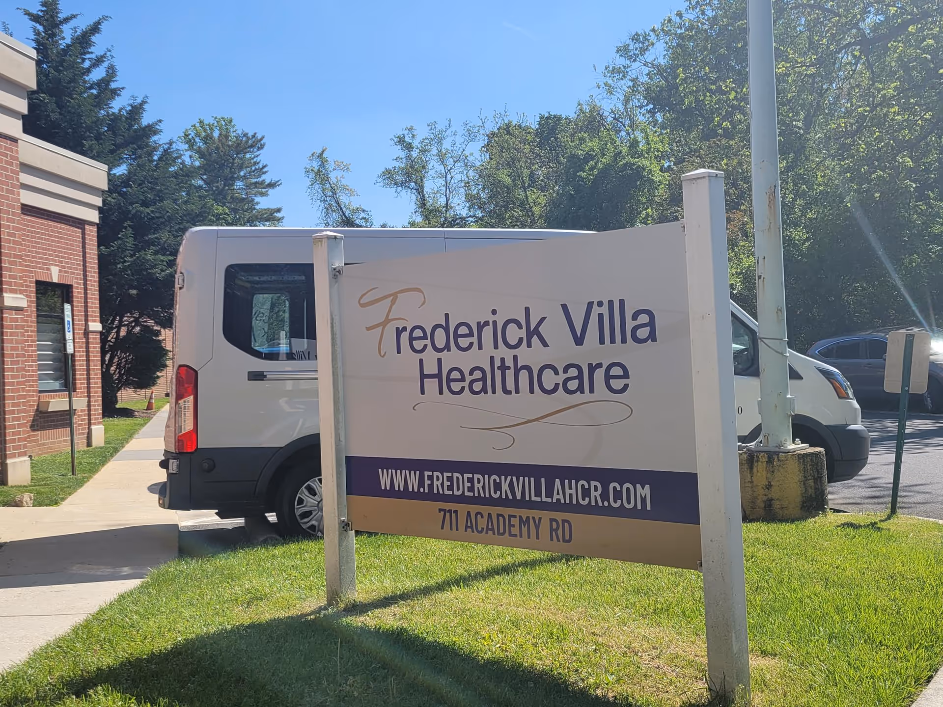 A freestanding sign on the lawn in front of a brick building reads "Frederick Villa Healthcare" with the website and address and a parked van behind it.