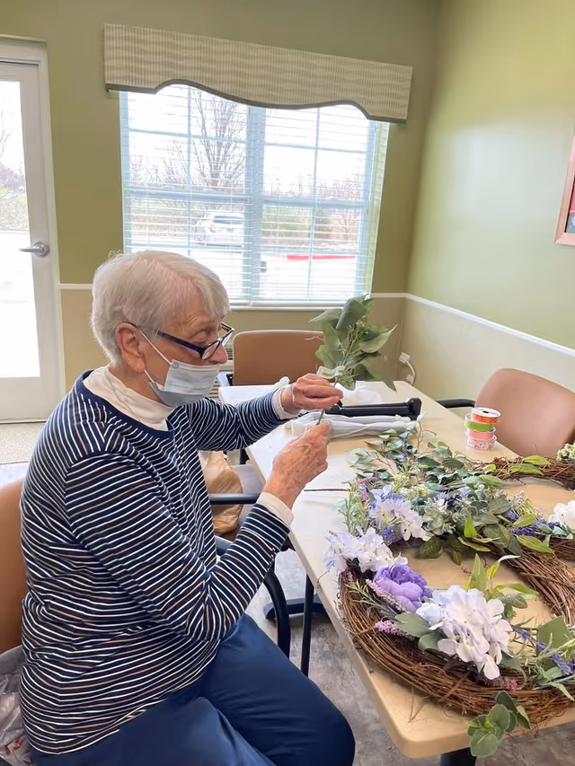 An elderly woman wearing a face mask and glasses is seated at a table in a well-lit room with green walls and a window. She is arranging artificial flowers and greenery on a wreath. Various craft supplies and floral materials are spread out on the table.