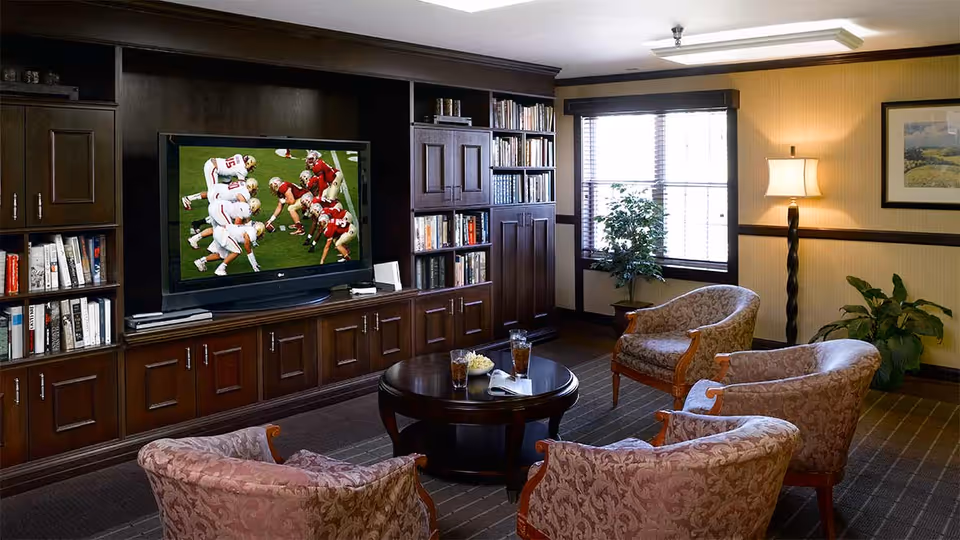 A cozy living room with four patterned armchairs arranged around a dark wooden round coffee table holding two glasses of iced tea and a bowl of popcorn. A large flat-screen TV mounted on a dark wood built-in cabinet is showing a football game. Bookshelves filled with books flank the TV. A window with blinds and two potted plants are visible in the background, along with a floor lamp and framed artwork on the wall.