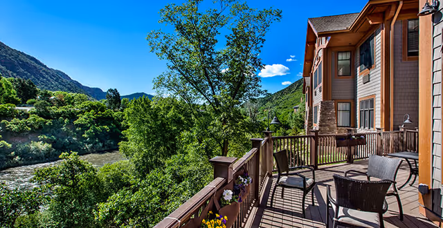A wooden balcony with outdoor chairs and tables overlooking a lush green landscape with trees and a river, adjacent to a multi-story building under a clear blue sky.