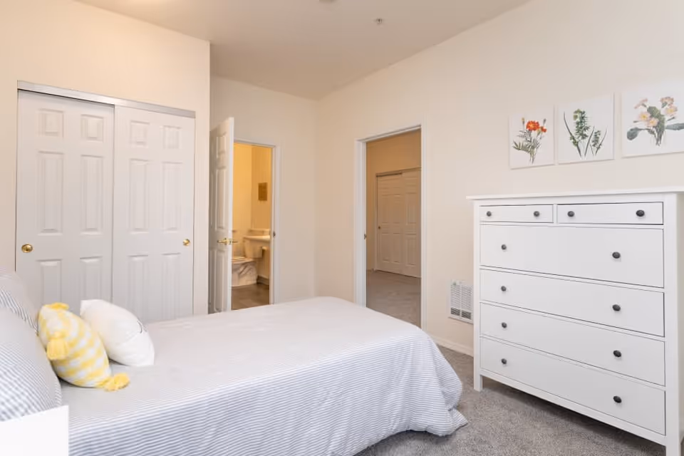 A bedroom with a bed covered in a light striped bedspread and two pillows, one yellow and one white. There is a white dresser with six drawers on the right side of the room. Three botanical prints hang on the wall above the dresser. Two doors are visible: one leading to a bathroom with a toilet and sink, and the other leading to another room with a closet. The walls are painted light beige and the floor is carpeted.