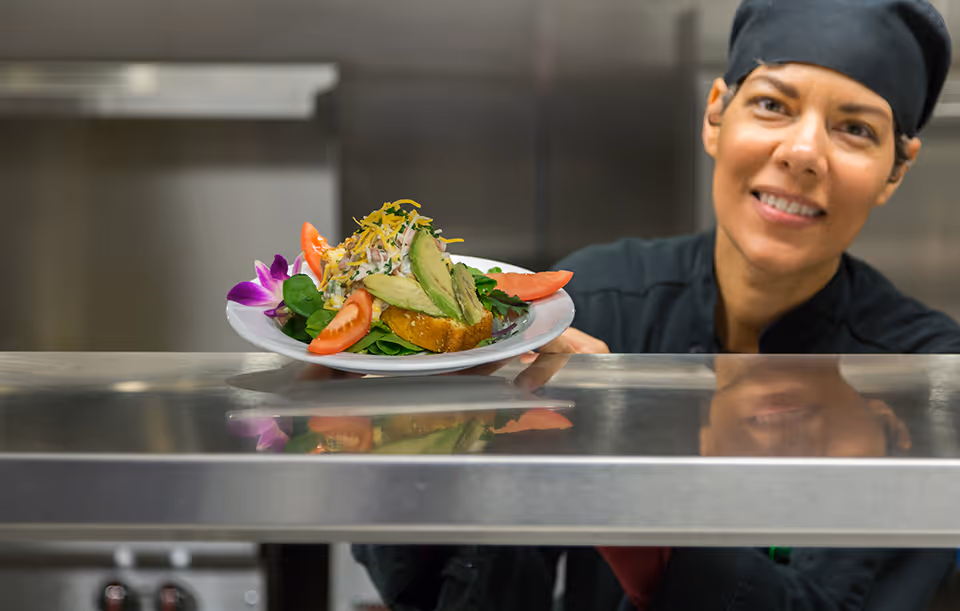 A chef wearing a black uniform and hat is smiling while holding a plate of food with avocado slices, tomato wedges, shredded cheese, and greens in a kitchen setting.