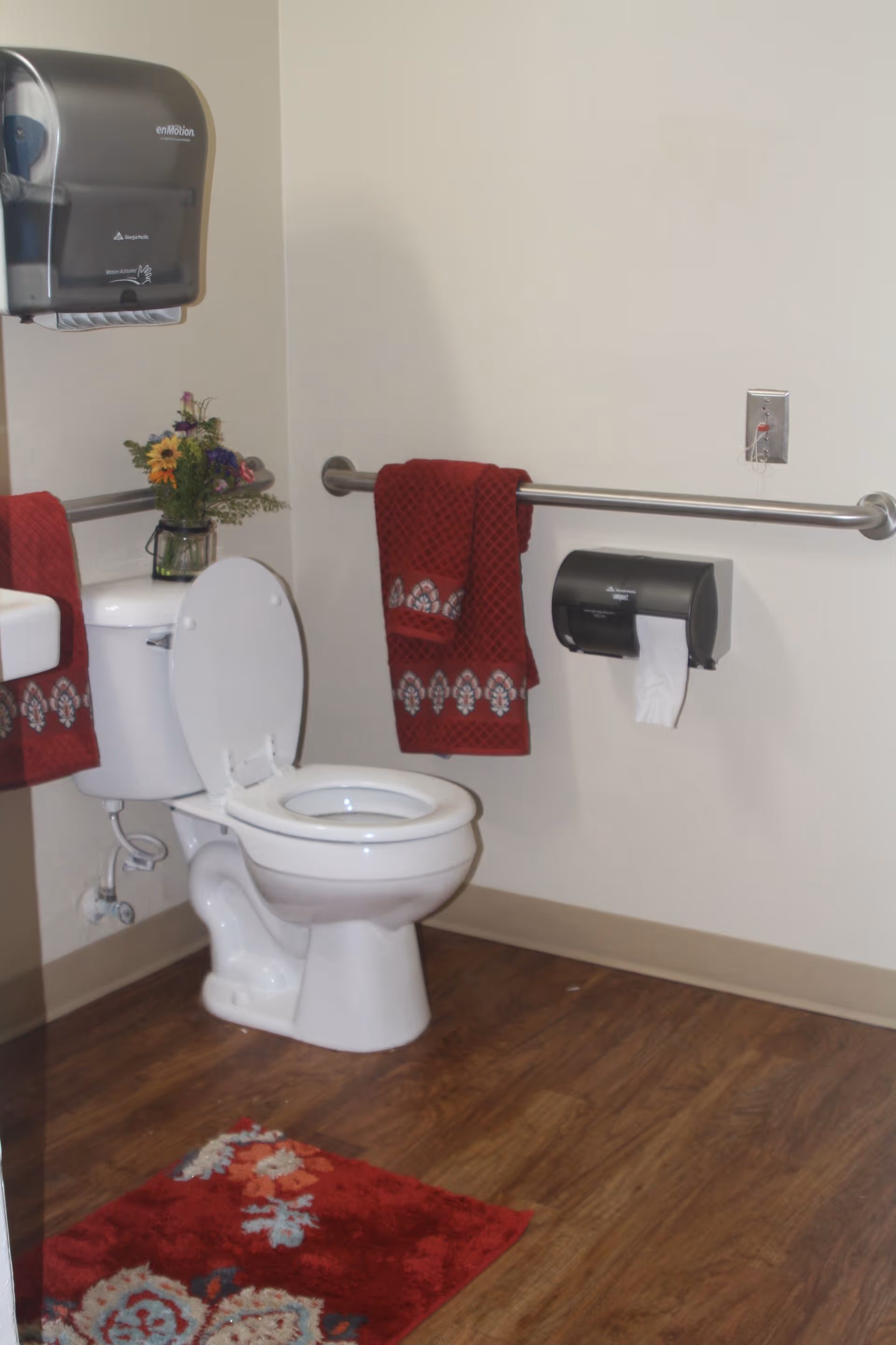 A clean bathroom with a white toilet, a red patterned rug on the wooden floor, red towels hanging on a metal grab bar, a wall-mounted paper towel dispenser, and a small vase with flowers on the toilet tank.