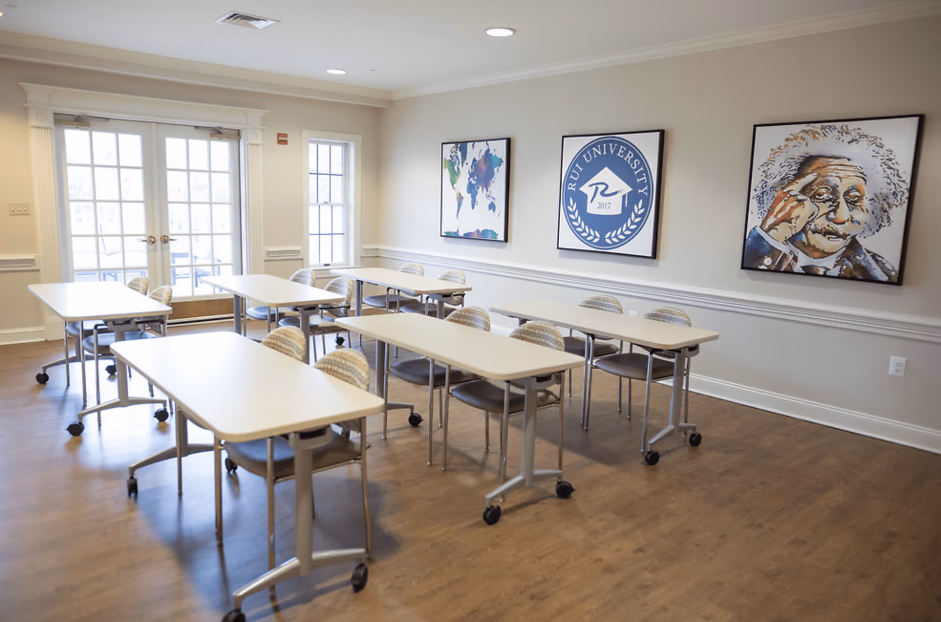 A bright room with several white tables and chairs arranged in rows, featuring wooden flooring and large windows with glass doors letting in natural light. The walls are decorated with three framed pictures, including a world map, a university emblem, and a portrait of Albert Einstein.