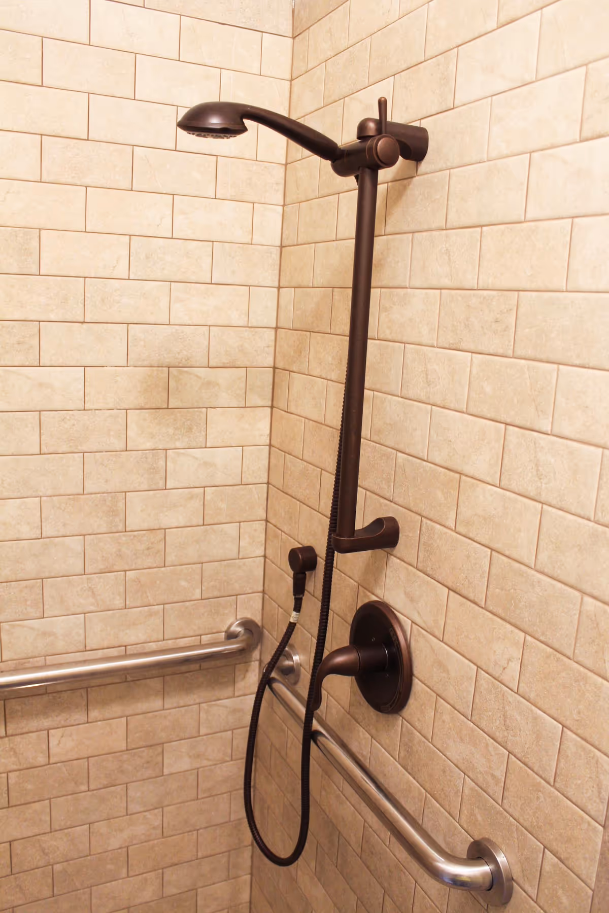 Shower area with beige tiled walls, an oil-rubbed bronze showerhead and handheld sprayer, and stainless steel grab bars.