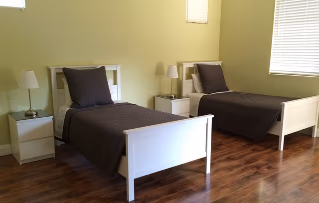 A simple bedroom with two single white beds, each with a dark gray blanket and pillow. Between the beds are two white nightstands, each with a small white lamp. The room has light green walls, wooden flooring, and two windows with blinds.