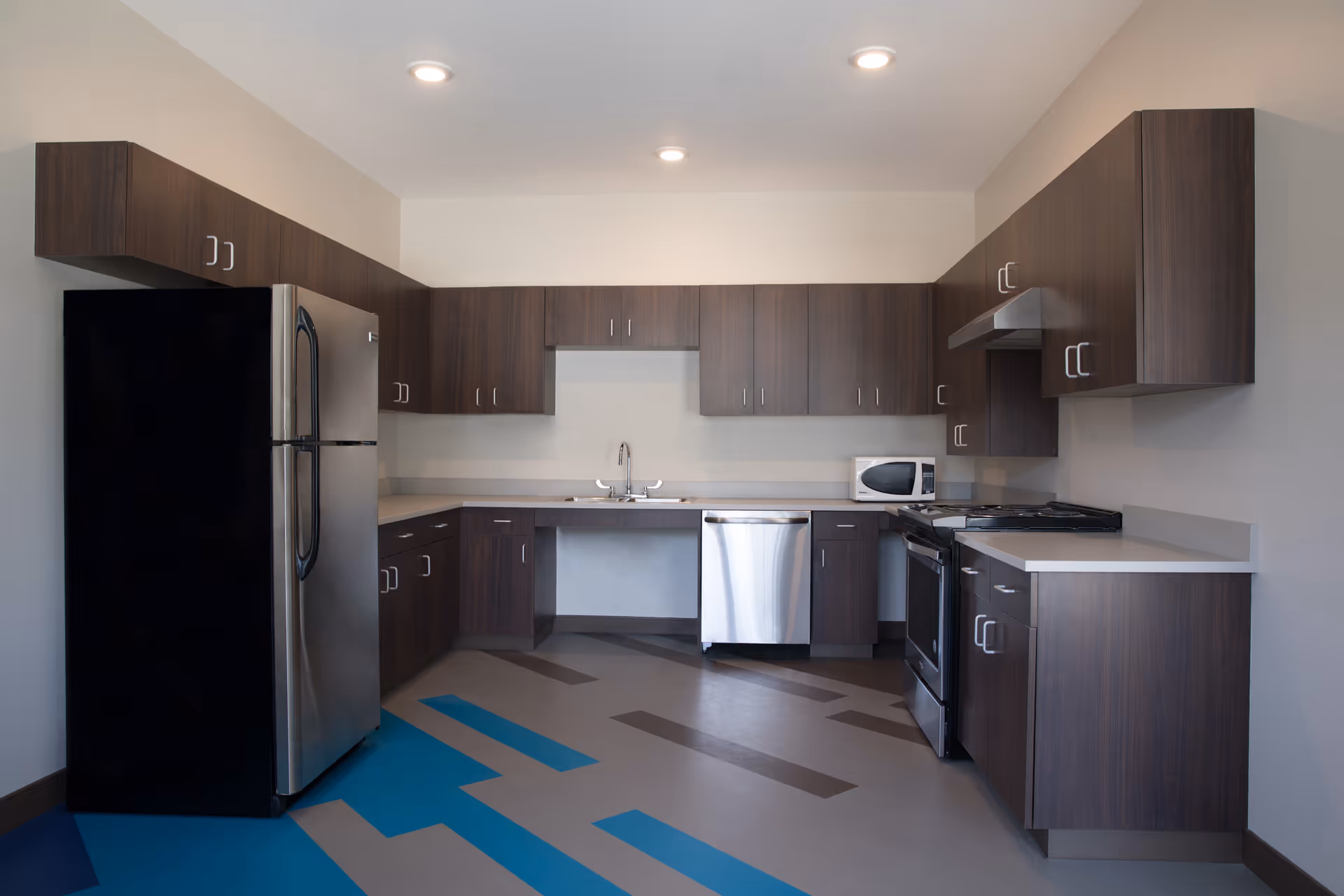 Modern kitchen with dark wood cabinets, stainless steel refrigerator, dishwasher, stove, microwave, and a sink under overhead recessed lighting. The floor has a geometric pattern with blue and gray colors.