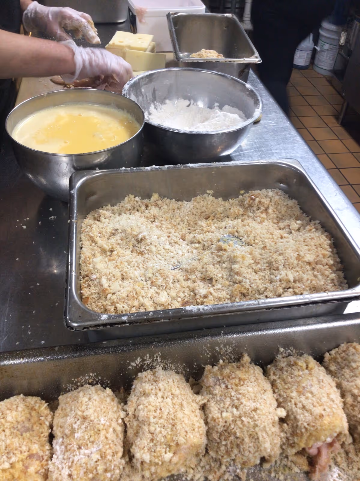 A commercial kitchen prep station with bowls of egg wash and flour and rows of breaded food being prepared on a stainless steel counter.