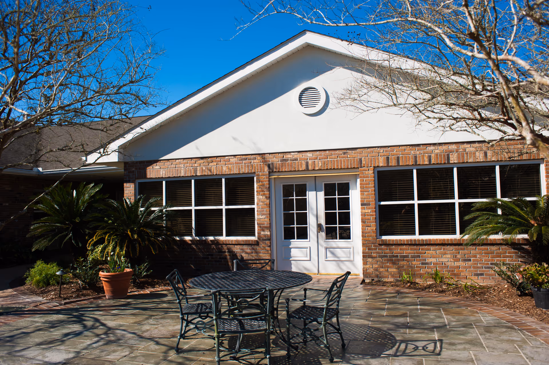 Brick building exterior with double glass doors, large windows, a patio table and chairs, and potted plants.