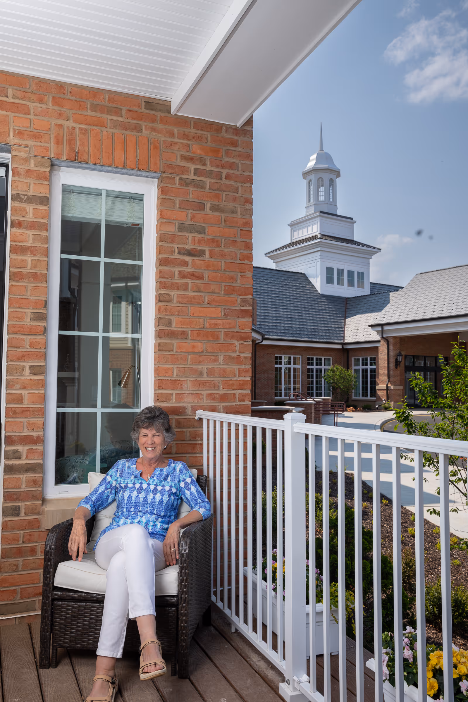 An elderly woman wearing a blue patterned blouse and white pants sits smiling on a cushioned wicker chair on a wooden porch. The porch has a white railing and is attached to a brick building. In the background, there is a courtyard with more brick buildings and a white cupola under a partly cloudy sky.