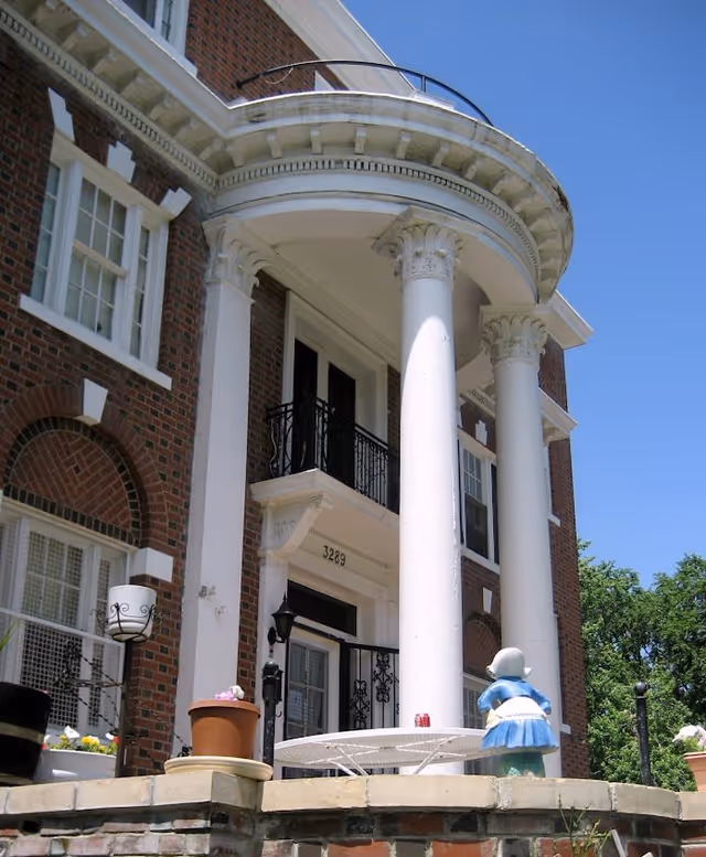 Front exterior view of a brick building with large white columns and a balcony above the entrance. There are potted plants and a small statue on a low brick wall in the foreground, with a clear blue sky and some trees in the background.