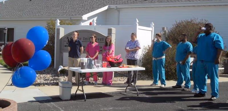 A group of seven healthcare workers standing outside near a table with refreshments including pitchers and cups. Four workers on the left wear pink and purple scrubs, while three on the right wear blue scrubs. There are red and blue balloons tied to a post on the left side. The background shows a white building and a sign partially visible behind the group.