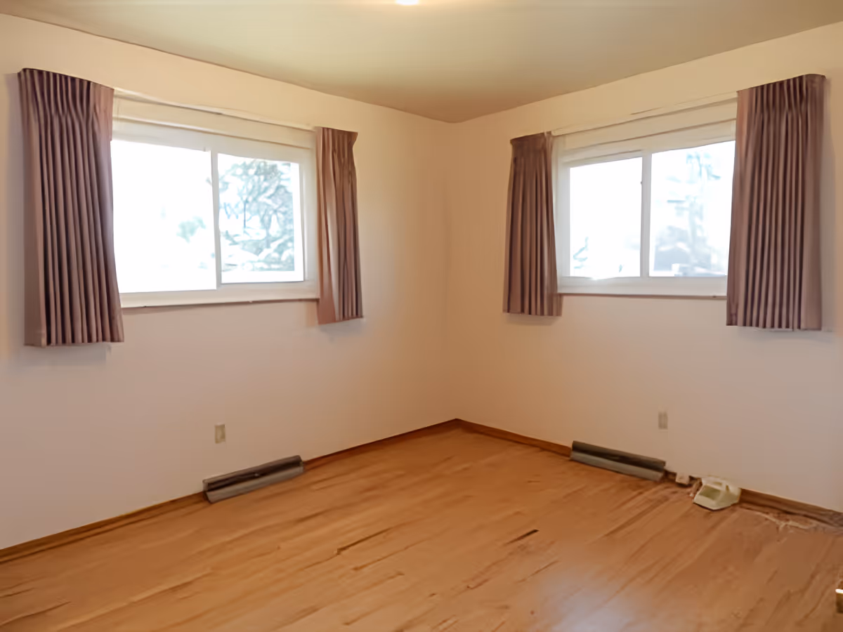 Empty room with two curtained windows and hardwood floors.