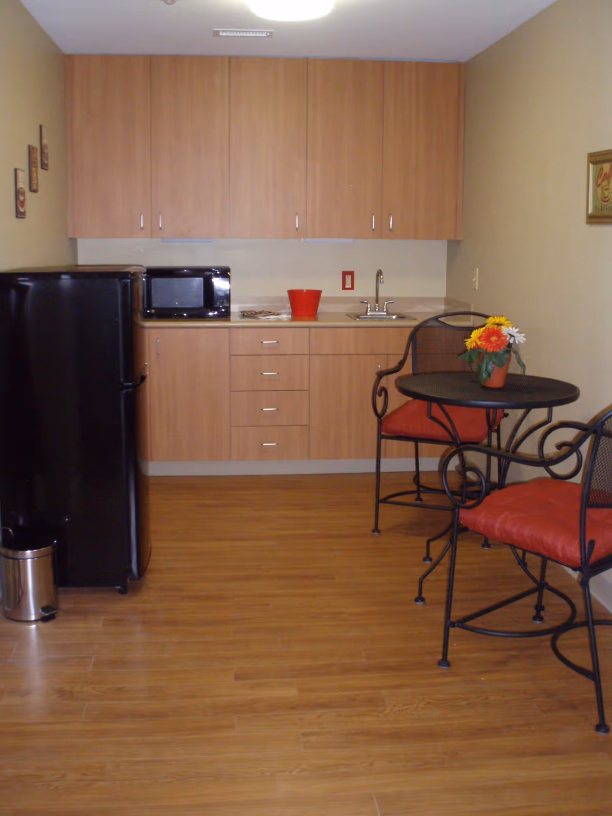Small kitchen area with light wood cabinets, a black refrigerator, a microwave, a sink, and a countertop. There is a small round table with two metal chairs that have red cushions. A small flower pot with colorful flowers is on the table. The floor is wood, and the walls are painted beige.