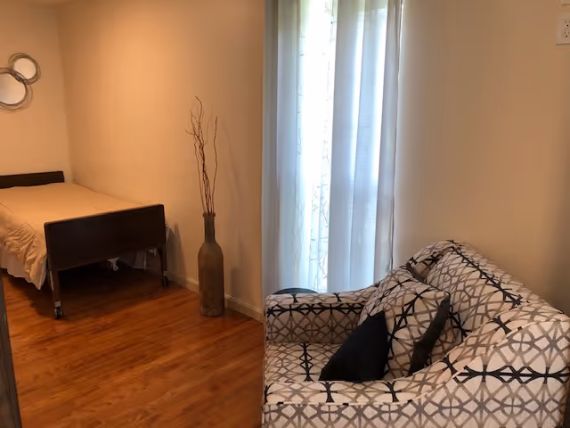 A room with a single bed with a beige bedspread on the left side, a tall decorative vase with dried branches next to the bed, and a patterned armchair with two dark cushions on the right side near a window with sheer white curtains.
