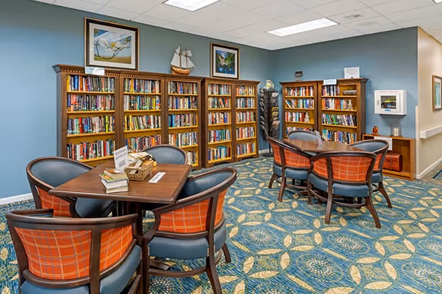 A cozy library room with multiple wooden bookshelves filled with books against blue walls. There are two wooden tables, each surrounded by four chairs with orange plaid upholstery. The carpet has a blue and beige floral pattern. The room is well-lit with ceiling lights and decorated with framed pictures and a model ship on top of the bookshelves.