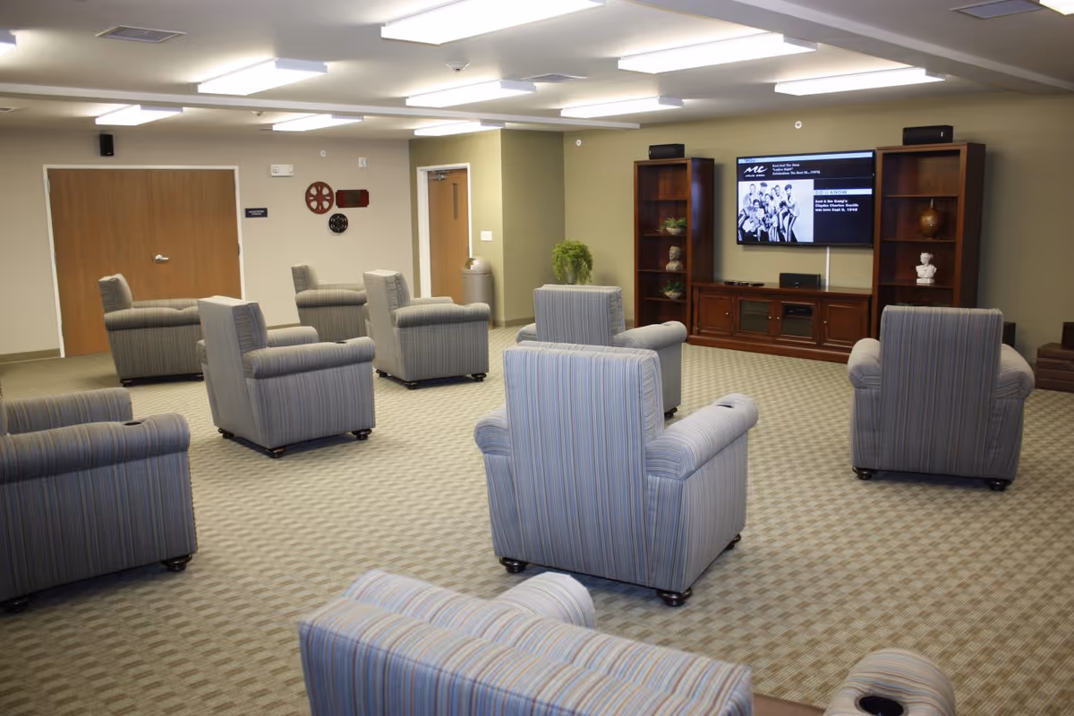 A common area with multiple striped armchairs arranged facing a wall-mounted flat screen TV. The room has beige walls, carpeted floor, wooden double doors, and wooden shelving units flanking the TV. The ceiling has multiple rectangular fluorescent light fixtures.
