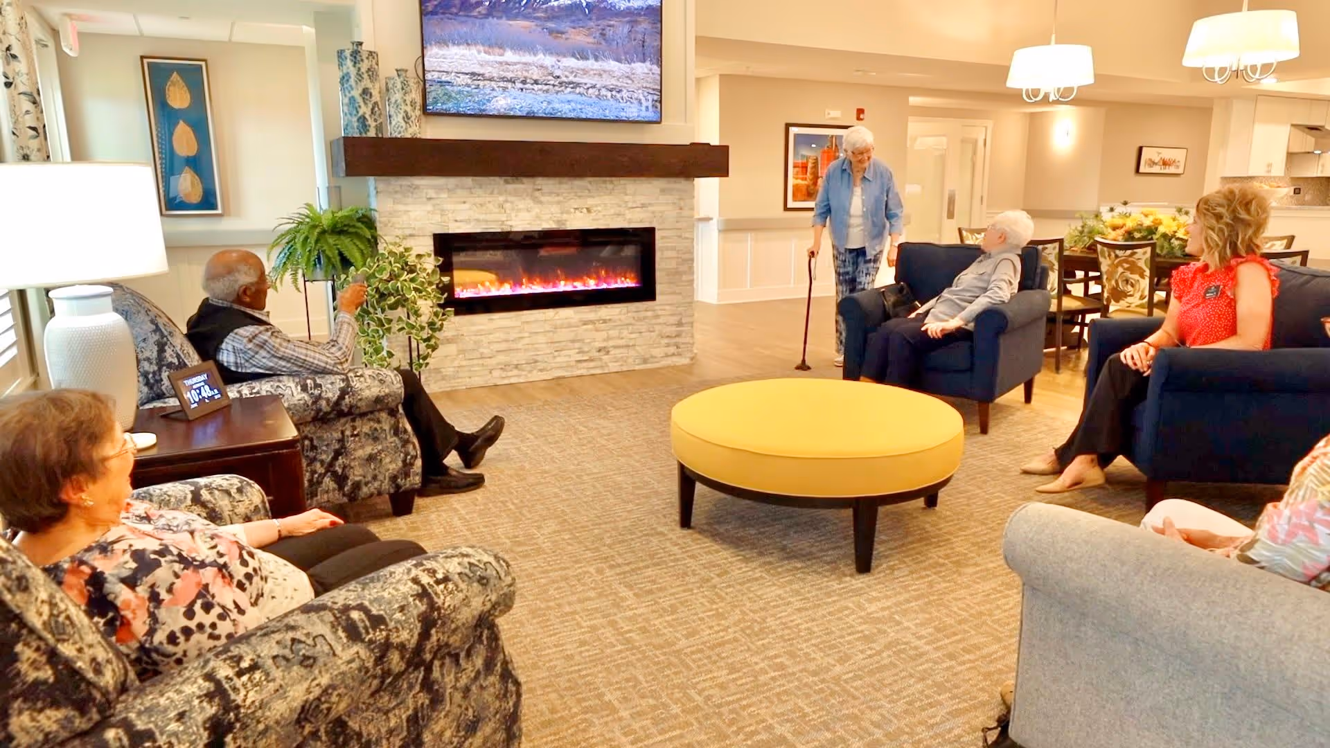 A cozy living room in an assisted living facility with several elderly residents and a staff member sitting and standing around a modern electric fireplace. The room features comfortable armchairs, a round yellow ottoman, a large TV mounted above the fireplace, decorative plants, and warm lighting.