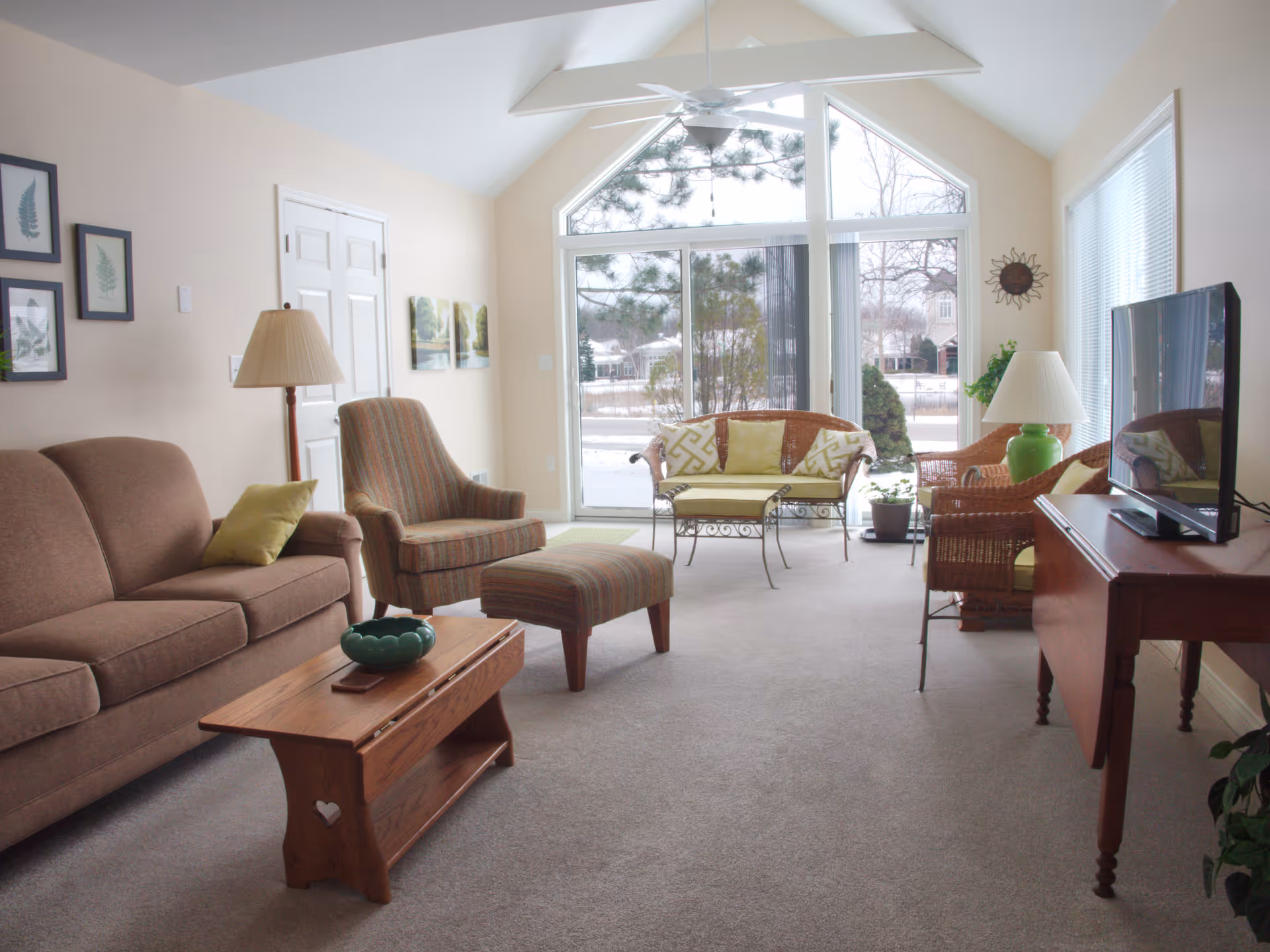 A bright living room with a large triangular window and sliding glass door showing an outdoor view. The room has a brown sofa with a green pillow, a striped armchair with matching ottoman, a wooden coffee table, a wicker loveseat with yellow cushions, and a wicker chair with a green lamp on a wooden side table. The walls are light-colored with framed artwork and a sun-shaped wall decoration.