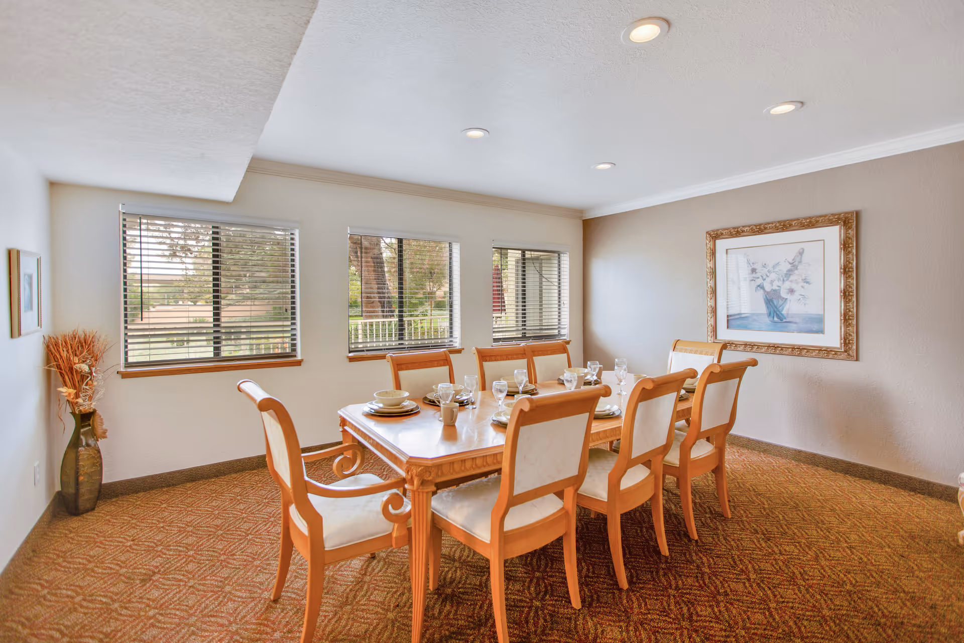 A dining room with a wooden dining table set for six people with plates, bowls, cups, and glasses. The room has three windows with blinds, a framed floral painting on the wall, a vase with dried flowers in the corner, and recessed ceiling lights.